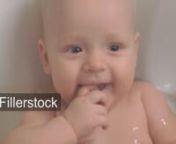 A closeup of a baby girl enjoying bathing. She is lying in babys bathtub in clear water, with some waterdrops on her face. A girl is smiling and laughing, putting her right hand in a mouthnLicense this clip: https://fillerstock.com/video/15996
